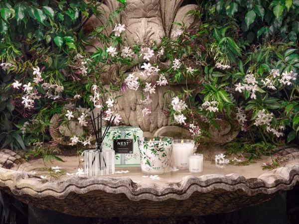 Still life of NEST New York candles and fragrance diffuser surrounded by jasmine flowers and lush greenery on a stone fountain.