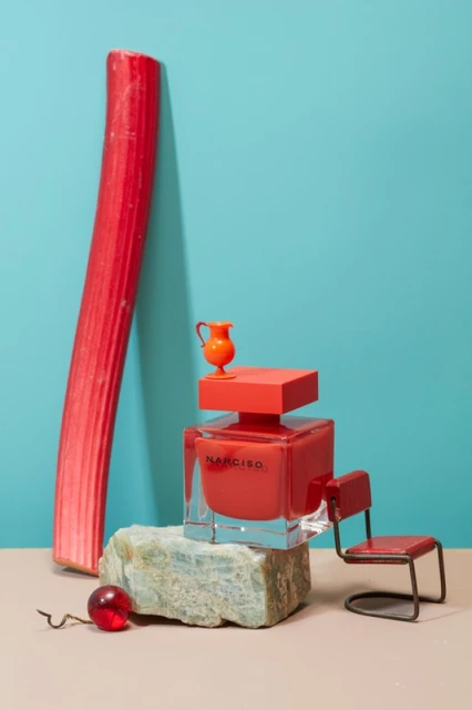 Still life of a bottle of red NARCISO perfume on a rock base, with rhubarb and a toy chair, set against a blue background.