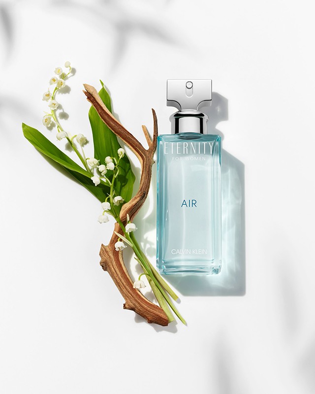 Calvin Klein Eternity Air perfume bottle stands with lily of the valley and a wood branch against a white surface with leaf shadows.