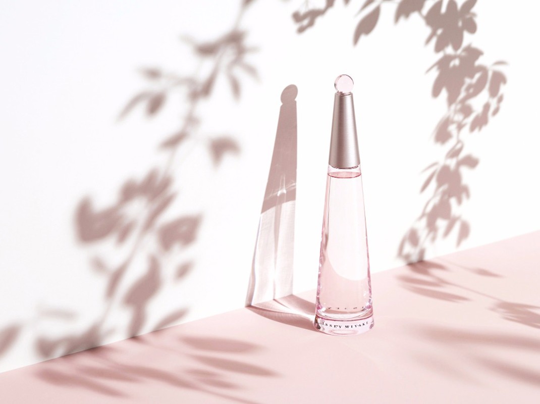 Perfume bottle on a pink surface against a white wall with soft floral shadows creating a minimalist and elegant composition.