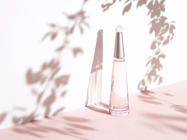 Perfume bottle on a pink surface against a white wall with soft floral shadows creating a minimalist and elegant composition.