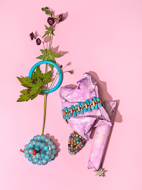 Still life with vintage jewelry: bead bracelets, turquoise belt against a pink background, flowers, and green leaves creating an artful composition.
