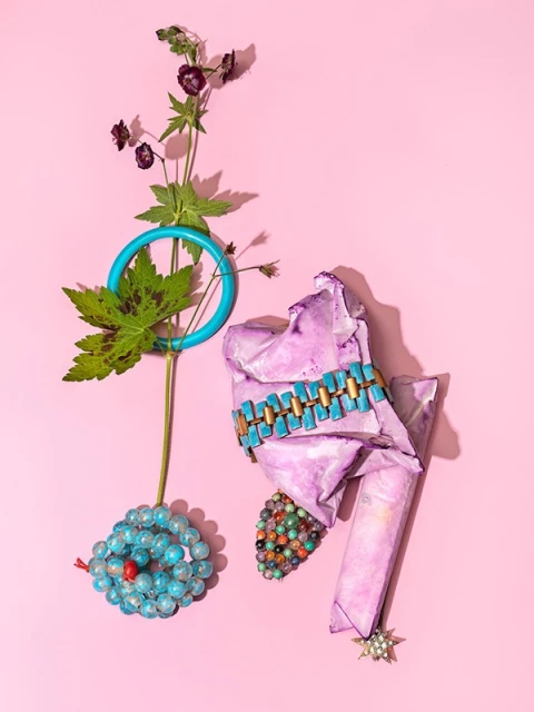 Still life with vintage jewelry: bead bracelets, turquoise belt against a pink background, flowers, and green leaves creating an artful composition.