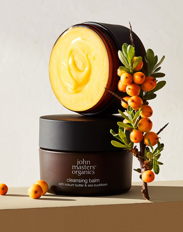 Two stacked brown jars of John Masters Organics cleansing balm with bright yellow product inside are decorated with sea buckthorn.