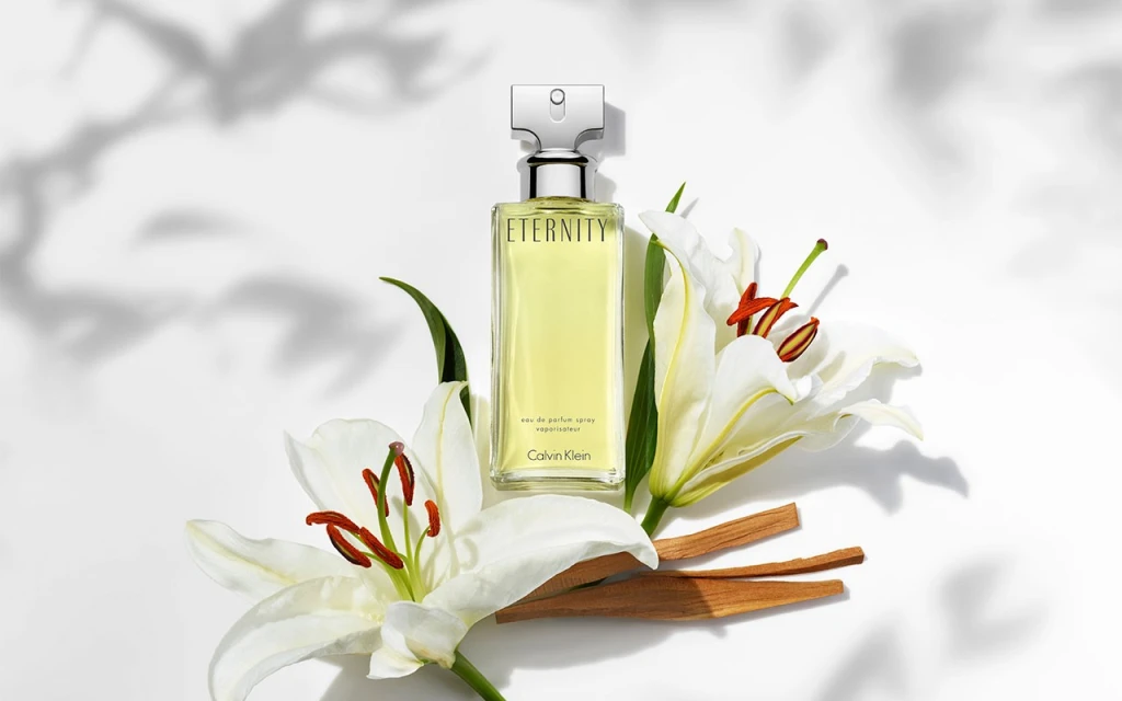 Eternity Calvin Klein perfume bottle on white surface with white lilies and cinnamon sticks, light yellow liquid inside the glass bottle.
