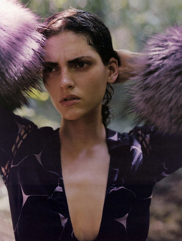 Portrait of an adult woman with wet hair and a printed, low-cut dress with fur accents, posing outdoors with a blurred, leafy background.