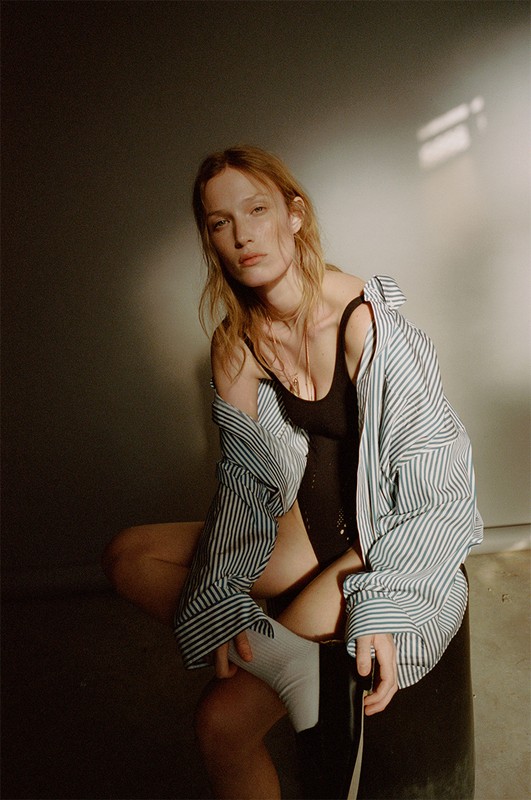 Portrait of a red-haired woman in a black bodysuit and striped shirt sitting indoors with soft lighting. Fashion and beauty photography.