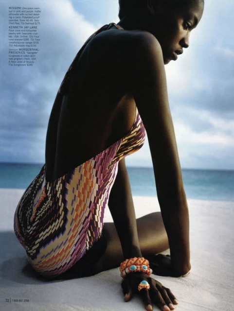 A woman in a patterned swimsuit sits on a white sand beach, accessorized with bracelets and a ring, with a cloudy sky backdrop.