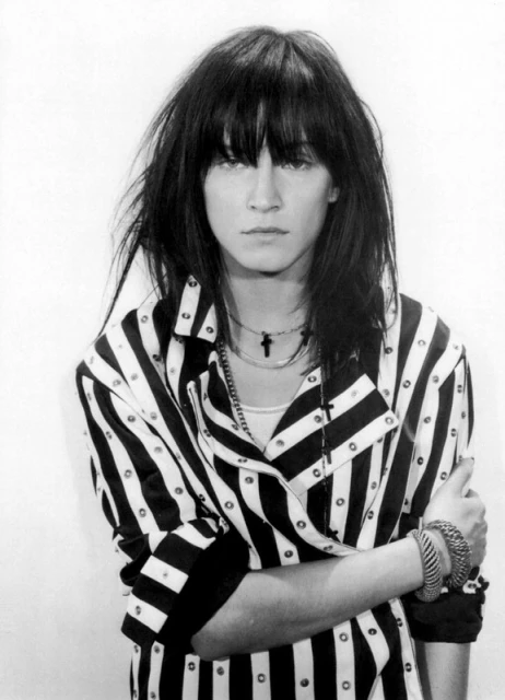 Black and white portrait of a woman with dark hair wearing a striped shirt and multiple necklaces posing indoors with folded arms.