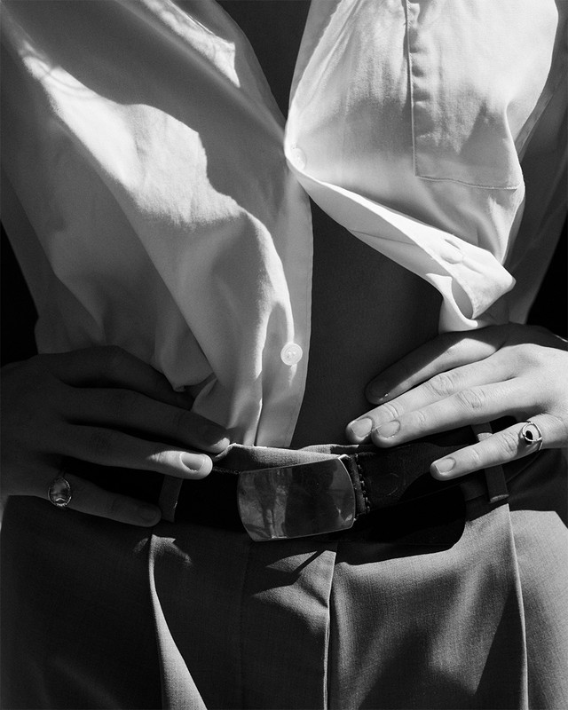 Close-up shot of a person's torso and hands wearing a white button-down shirt, belted pants, and rings, captured in black and white.