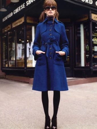 Adult woman wearing a blue coat and heart-shaped sunglasses standing in front of a store with an awning in an urban setting.