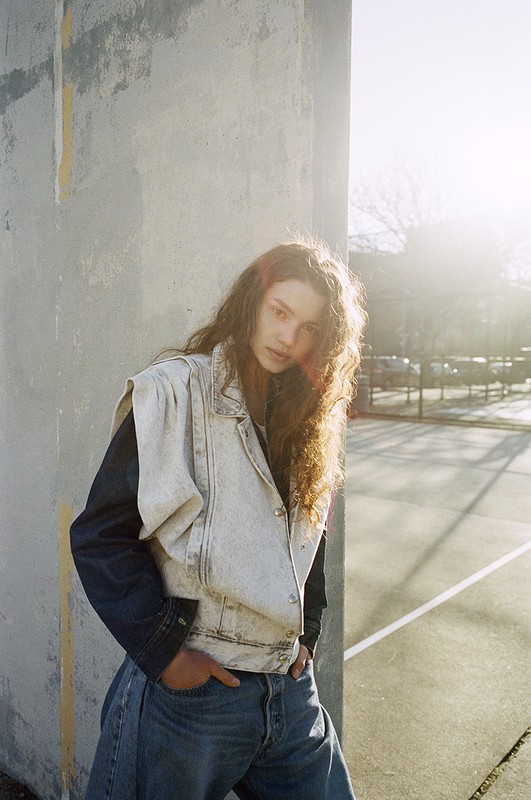 Woman with long curly hair in a sleeveless jean jacket and jeans poses near a concrete wall on a tennis court on a sunny day.