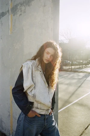 Woman with long curly hair in a sleeveless jean jacket and jeans poses near a concrete wall on a tennis court on a sunny day.