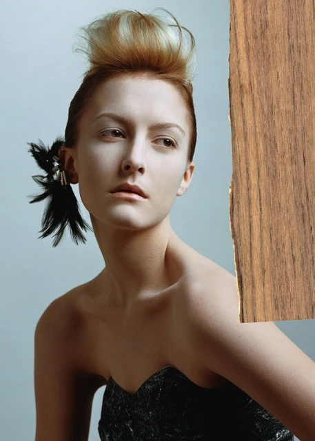 Portrait of a blonde woman with unique hairstyle and feather embellishment posing with wood panel. Beauty and fashion shot.
