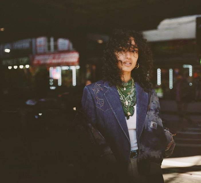Portrait of a young woman with curly hair in a denim jacket, wearing a green bandana necklace and hoop earrings in an urban setting.