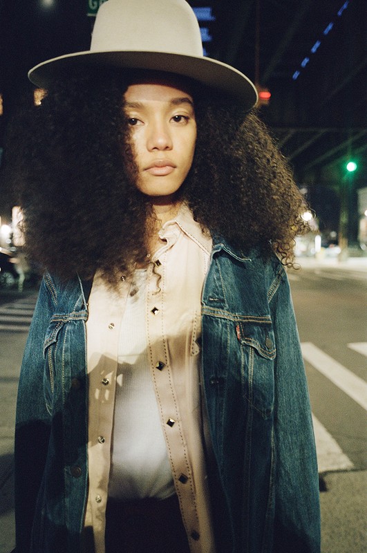 Stylish portrait of a woman with large curly hair wearing a hat and jean jacket standing on a street at night with soft, moody lighting.
