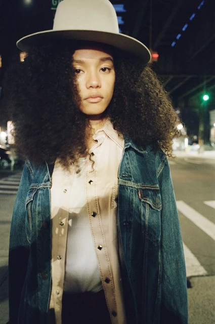 Stylish portrait of a woman with large curly hair wearing a hat and jean jacket standing on a street at night with soft, moody lighting.