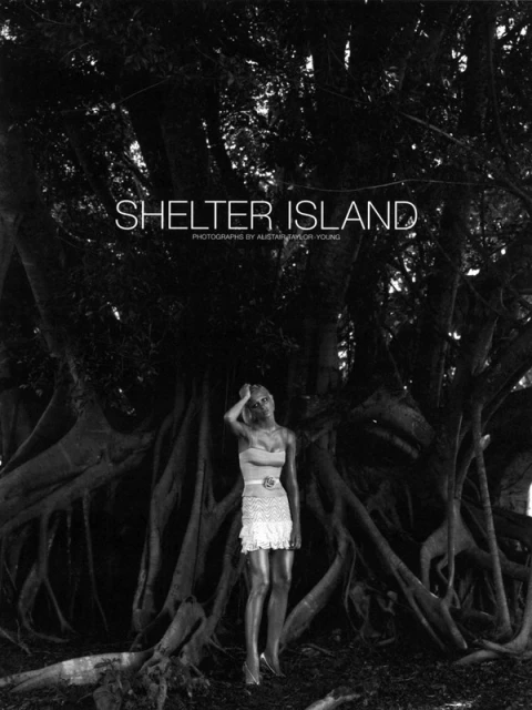Black and white image of a female model in a dress standing in front of a large tree with exposed roots, Shelter Island text overlay.