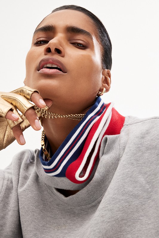 Close-up of a woman wearing a gray sweatshirt and a gold chain, and a unique red, white, and blue neckline, with gold gloves.