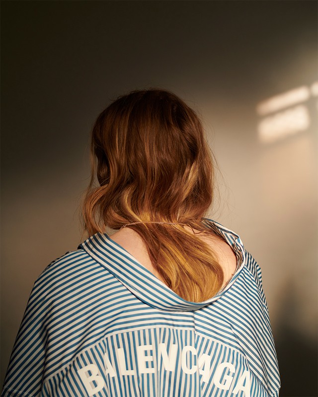 Back view of a young woman with reddish-brown hair wearing a blue and white striped Balenciaga shirt in an indoor setting.