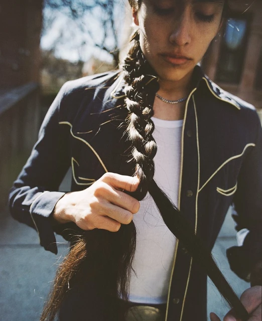 Young adult with a long braid and wearing a navy jacket with gold piping stands outside. Styling her hair in natural daylight.
