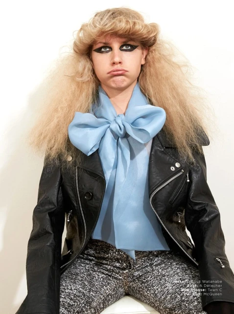 Teen girl with extreme eye makeup and blonde crimped hair wears a black leather jacket and blue bow blouse against a white wall.