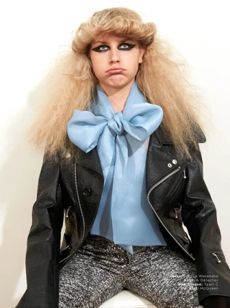 Teen girl with extreme eye makeup and blonde crimped hair wears a black leather jacket and blue bow blouse against a white wall.