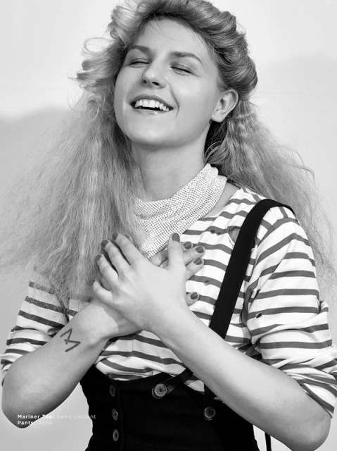 Black-and-white portrait of an adult female smiling with hands clasped to her chest, wearing striped shirt and dark bib overalls.