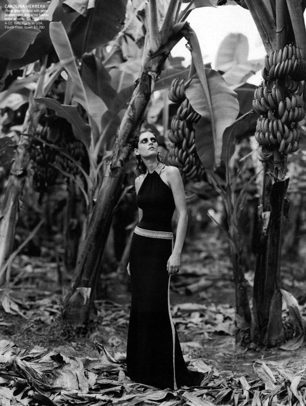 A model in a black gown poses in a banana plantation with bunches of bananas hanging from the trees in this black-and-white fashion shot.