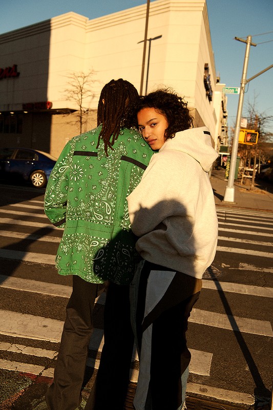 Two people stand on a crosswalk in an urban setting. A woman looks towards the camera. The man's back is to the camera.