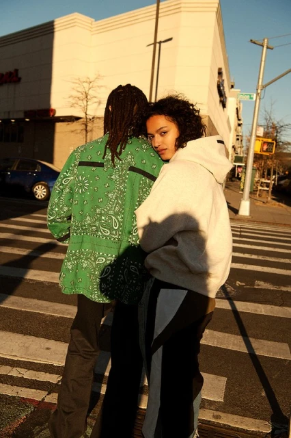 Two people stand on a crosswalk in an urban setting. A woman looks towards the camera. The man's back is to the camera.