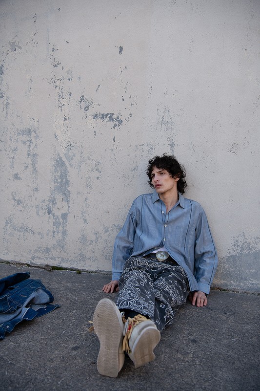 Man in a blue button-down shirt and patterned pants sitting against a weathered wall outdoors, wearing moccasins and a belt buckle.