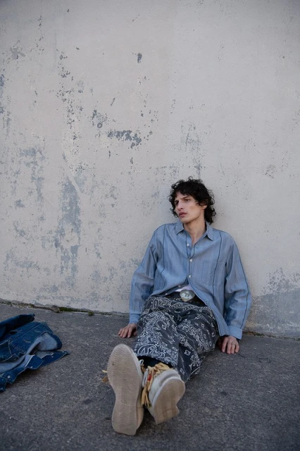 Man in a blue button-down shirt and patterned pants sitting against a weathered wall outdoors, wearing moccasins and a belt buckle.