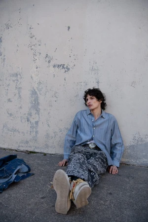 Man in a blue button-down shirt and patterned pants sitting against a weathered wall outdoors, wearing moccasins and a belt buckle.