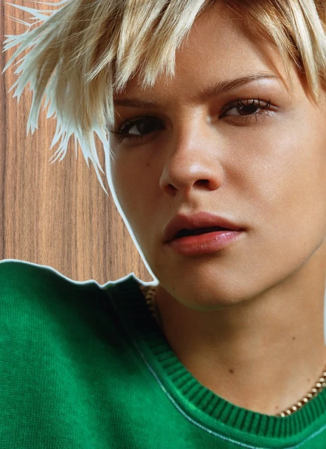 Close-up portrait of a young adult female with short blonde hair and a green sweatshirt against a wood-grain background.