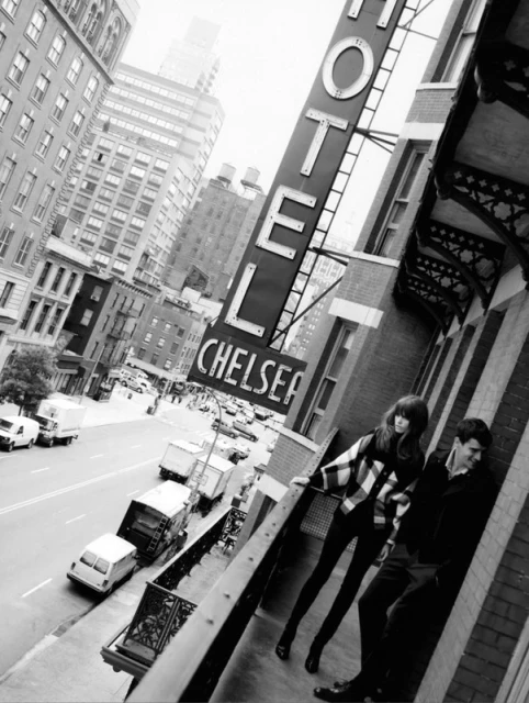 Black and white image of a couple on a hotel balcony, with the Chelsea Hotel sign above and city buildings in the background.