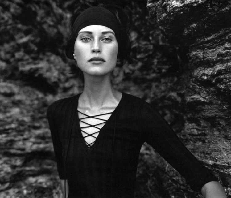 Black and white portrait of a woman with a headscarf wearing a V-neck top, posing against a dark, textured rock wall.