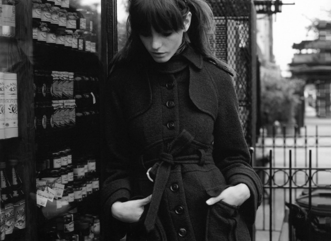 A woman in a stylish overcoat stands in front of a store window displaying jars and bottles in a black-and-white outdoor scene.