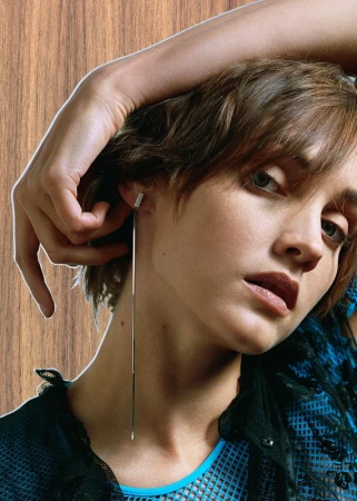 Close-up portrait of a woman with short brown hair wearing a long silver drop earring and a blue mesh top against a wood-textured background