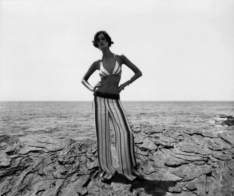 A female model in a striped bikini top and wide-leg pants poses on a rocky shore with the ocean and clear sky in the background.