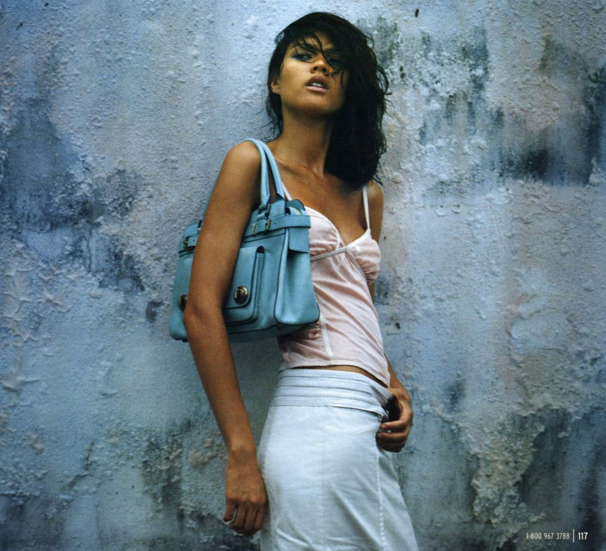 Woman in pink top and white skirt carrying a blue handbag against a distressed wall by Lorri Sendel