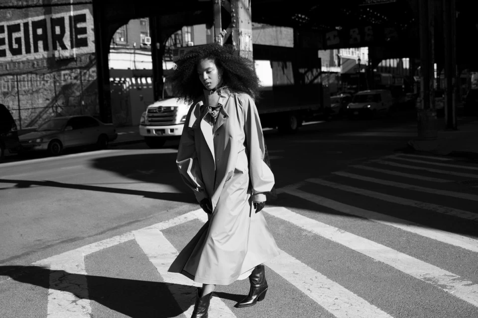 A monochrome fashion shot of a woman with large hair crossing a zebra striped crosswalk in a long coat and boots in an urban setting.