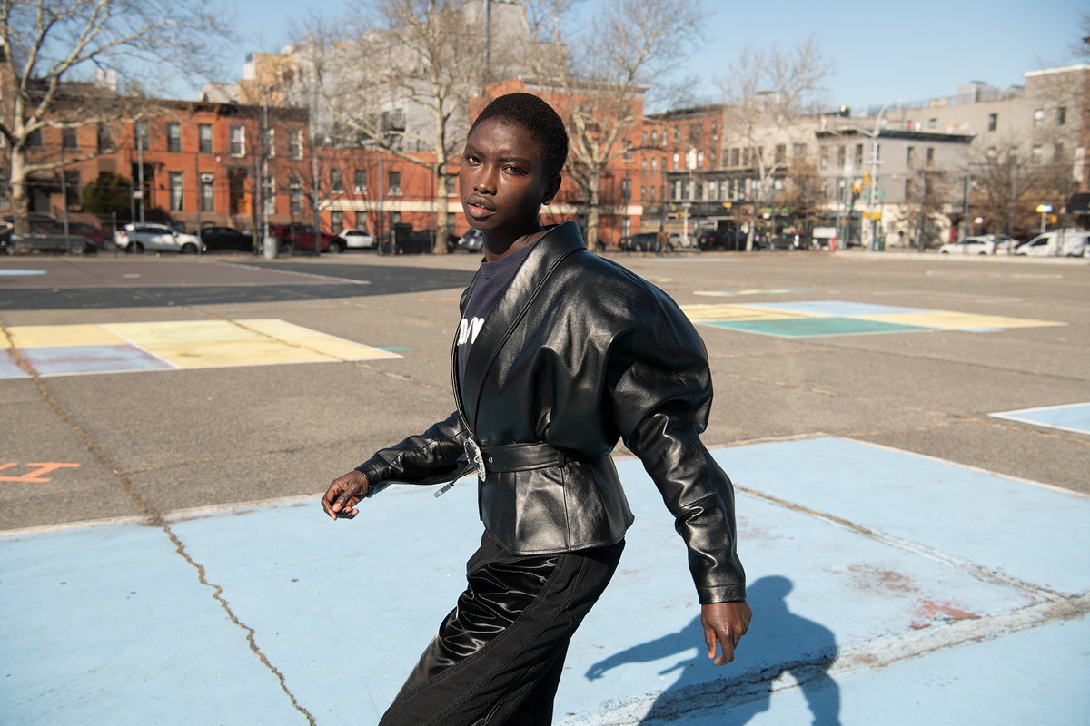 A slender woman with dark skin strides confidently across a painted urban court, wearing a black leather jacket and shiny black pants.