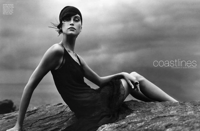Monochrome fashion shot of a woman in a flapper-style dress sitting on rocks, set against a cloudy sky with coastlines text overlay.