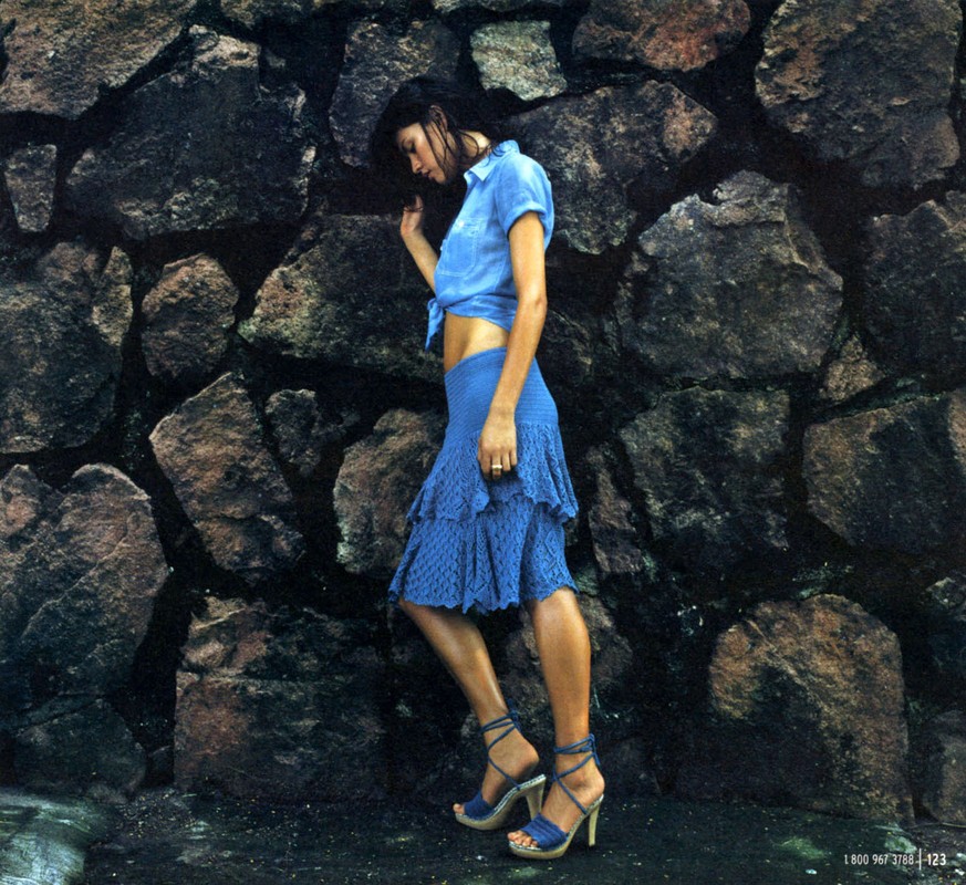 Brunette woman in blue top and skirt poses against a textured stone wall with high-heeled sandals, fashion shot with a natural feel.
