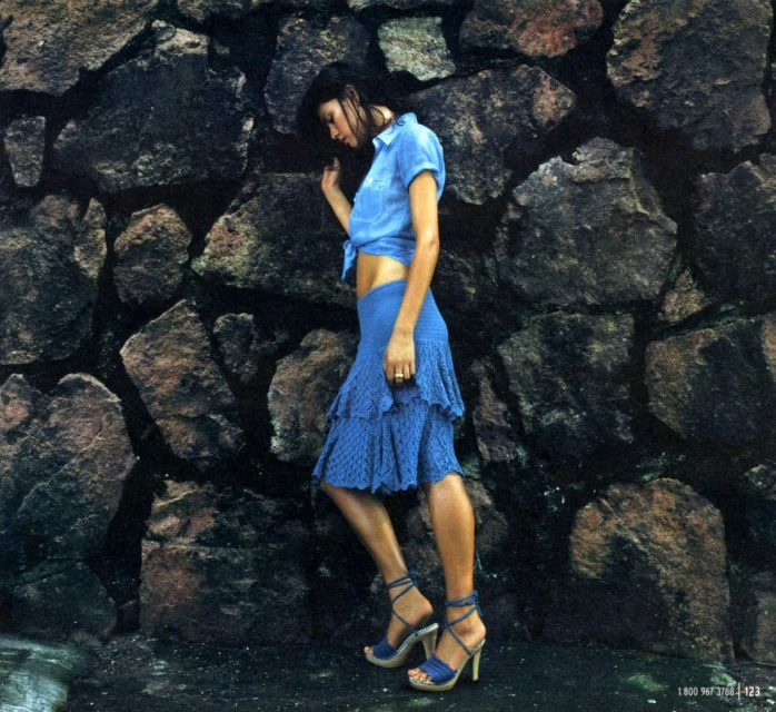 Brunette woman in blue top and skirt poses against a textured stone wall with high-heeled sandals, fashion shot with a natural feel.