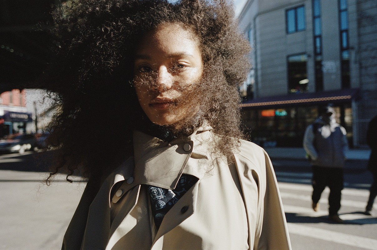 Close-up of a woman with curly hair in an urban setting. She is wearing a tan coat. Pedestrians cross a street in the background.