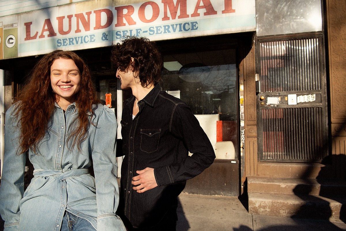 A smiling woman in denim stands beside a man in black outside a laundromat on a city street, bathed in natural light.