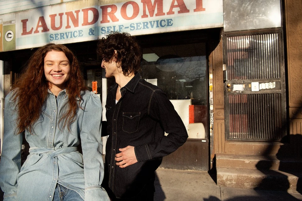A smiling woman in denim stands beside a man in black outside a laundromat on a city street, bathed in natural light.