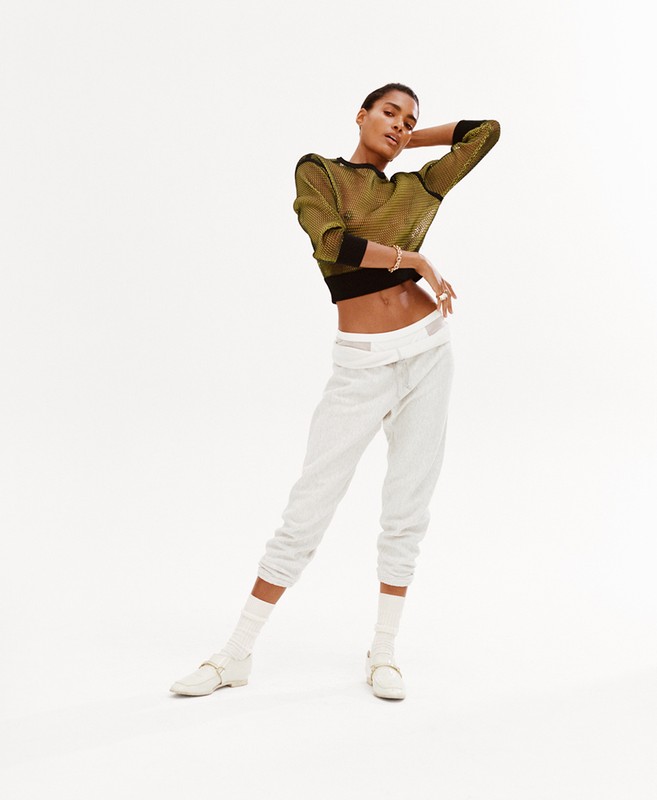 Fashion photo of a young Black female model in a sheer olive-green top and white sweatpants, striking a pose in a bright studio.